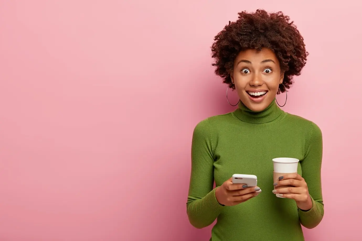Радостная кудрявая женщина со здоровой кожей в повседневной зеленой водолазке держит современный мобильный телефон и кофе навынос, наслаждается онлайн-общением.