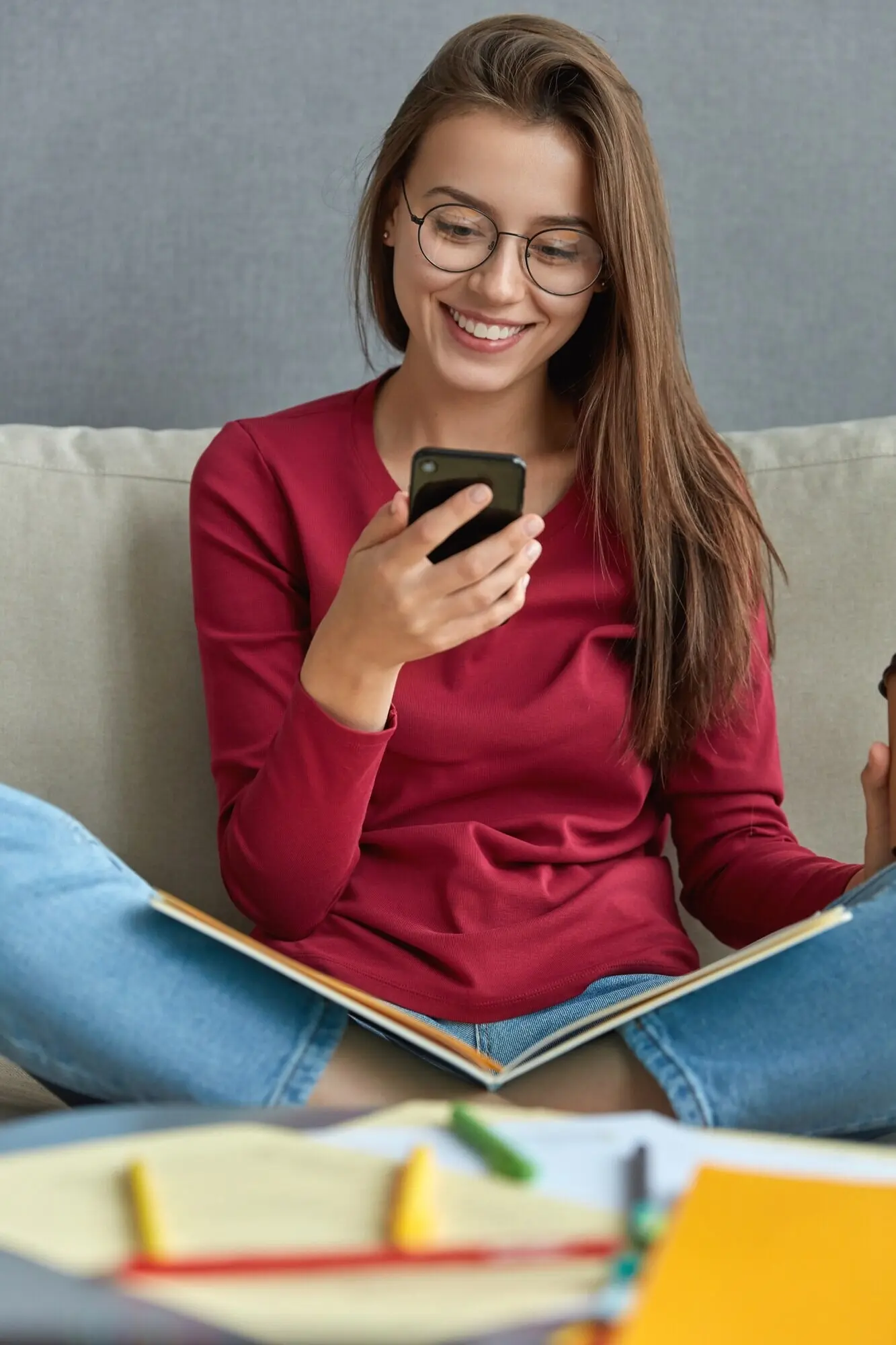 Довольная милая брюнетка обновляет профиль в социальных сетях, держит телефон, устанавливает приложение на цифровой сотовый телефон, сидит на диване, скрестив ноги, с книгой на коленях; на переднем плане — стол с бумагами и ручками.